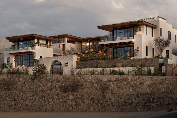 Exterior view of a large multi-level residential complex on a terraced, landscaped site. The white stucco buildings feature broad timber-toned projecting roof eaves, extensive floor-to-ceiling glazing, balconies, warm exterior lighting, and a prominent rough stone retaining wall. The architecture has a refined contemporary Mediterranean character under an overcast evening sky.