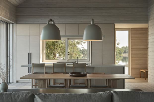 A refined open-plan kitchen and dining area viewed from behind a sofa. The room features a long island with a dark benchtop, integrated cabinetry in pale grey-beige tones, a timber dining table with upholstered chairs, twin oversized pendant lights, and large windows overlooking greenery and water. Timber boards cover the ceiling and portions of the walls, while full-height glazing and a light, muted palette create a calm connection to the outdoors.