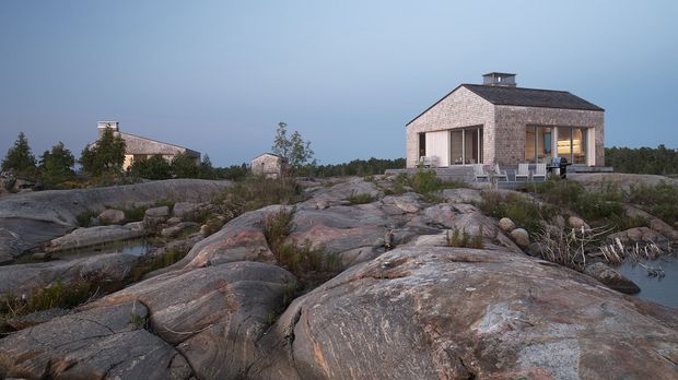 Exterior view of a compact modern house set among exposed granite bedrock and sparse natural vegetation. The main building has a simple rectangular form, a low pitched dark roof with a small chimney, pale textured masonry cladding, and large glazed openings. Additional small structures are visible in the background, with a rocky shoreline or water inlet at the right.