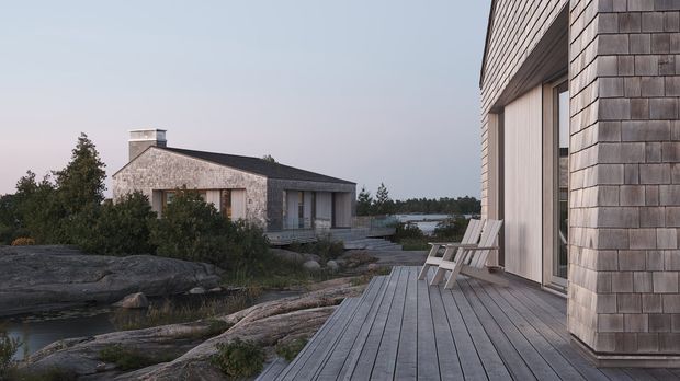 A contemporary coastal residence set among exposed rocky terrain and low vegetation. The foreground shows a broad timber deck with simple outdoor chairs, large sliding glazed doors, and a shingle-clad facade. A second low, dark-roofed dwelling is visible beyond, with water and a wooded horizon in the background. The scene has a restrained, natural palette and calm dusk atmosphere.