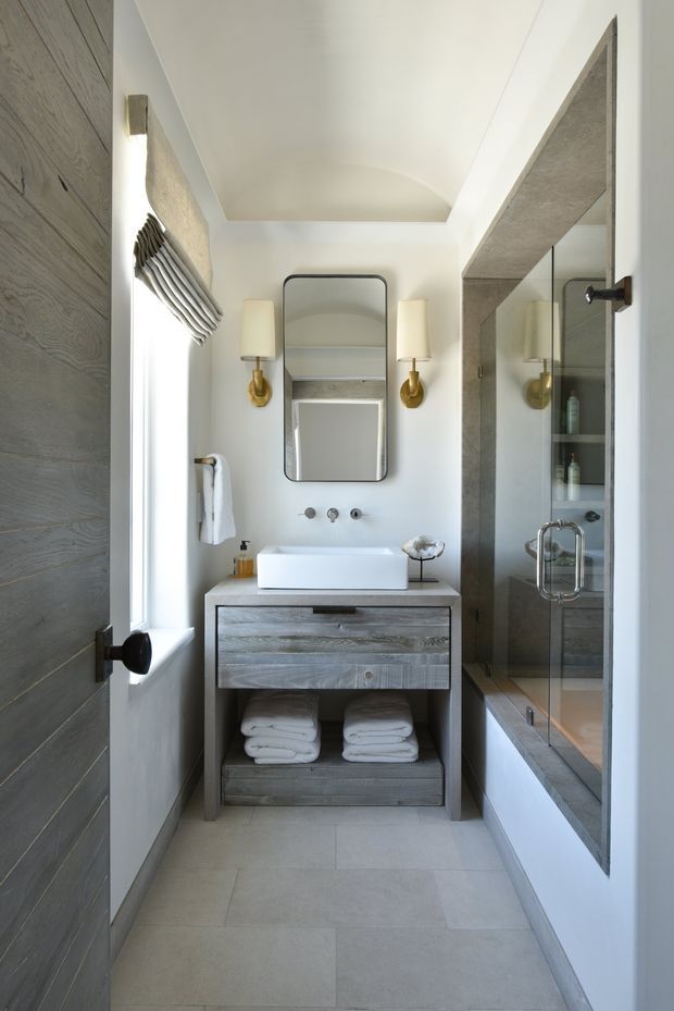 A narrow, refined bathroom with a floating-style grey wood vanity, white vessel basin, wall-mounted tapware, brass wall sconces, and a mirrored medicine cabinet. Large pale floor tiles and grey stone-look wall surfaces frame a glass shower enclosure beside a built-in bathtub. A timber door and textured wood joinery add warmth to the otherwise light, neutral space.