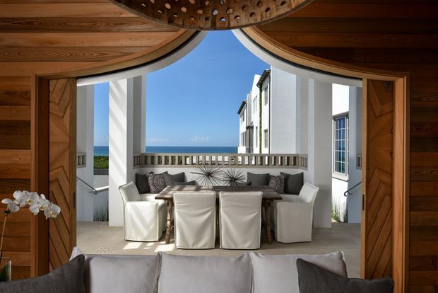 A framed interior view through an ornate timber opening toward a bright coastal dining and lounge area. The foreground shows the back of a light sofa, while the central space contains a dining table with upholstered slipcovered chairs, flanked by white architectural columns and open timber doors. Beyond the balustrade, the ocean is visible. The space combines warm wood detailing with white walls, abundant daylight, and a refined coastal character.