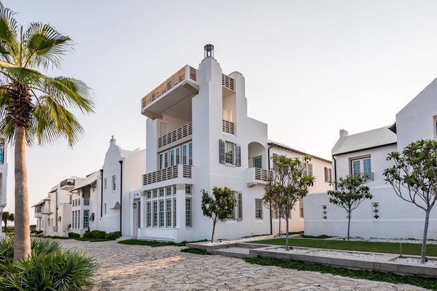 A bright exterior view of a white multi-storey residential development with arched openings, balconies, decorative railings, tall windows, and pitched metal roofs. The buildings are arranged along a landscaped paved walkway, with palm trees and tropical planting contributing to a resort-like setting.
