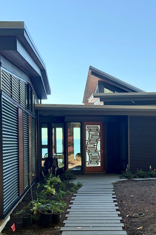 Exterior view of a contemporary coastal residence viewed from a landscaped entry path. The home has dark cladding, strong horizontal roof planes, large glazed doors and windows, external louvred shutters, and a prominent timber entry door with a geometric decorative panel. Water and distant shoreline are visible through the entry glazing. The composition highlights the covered entry courtyard and layered flat and pitched roof forms.