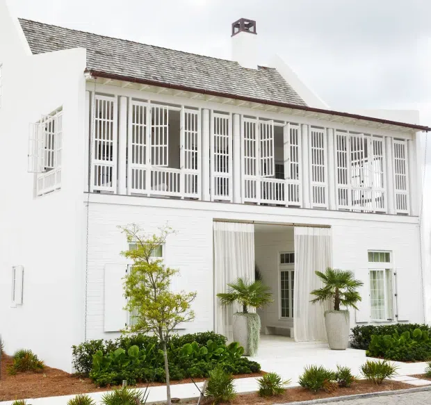 White two-storey residential facade with a long upper-level veranda enclosed by ornate vertical slatted screens, a weathered pitched shingle roof, and a prominent central entry opening framed by sheer curtains. Tropical palms, clipped hedges, and structured landscaping line the bright white exterior.