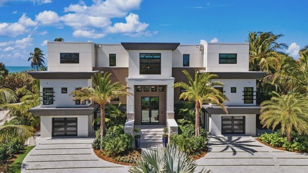 Front exterior view of a large symmetrical contemporary multi-storey residence, with white rendered geometric volumes, dark horizontal roof overhangs, broad glazing, two integrated garages, and a central recessed entry. The landscaped frontage includes palms and tropical planting, with blue sky and a glimpse of water or horizon behind the property.
