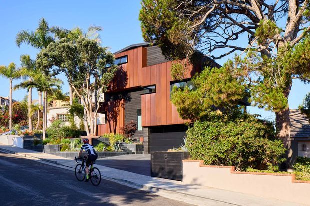 Street-level exterior view of a contemporary multi-storey residence with a strongly articulated facade combining warm reddish-brown vertical cladding, dark horizontal panels, and recessed horizontal glazing. The house sits behind landscaped front garden walls and a driveway in a leafy suburban setting, with mature trees and palms partially screening the building.