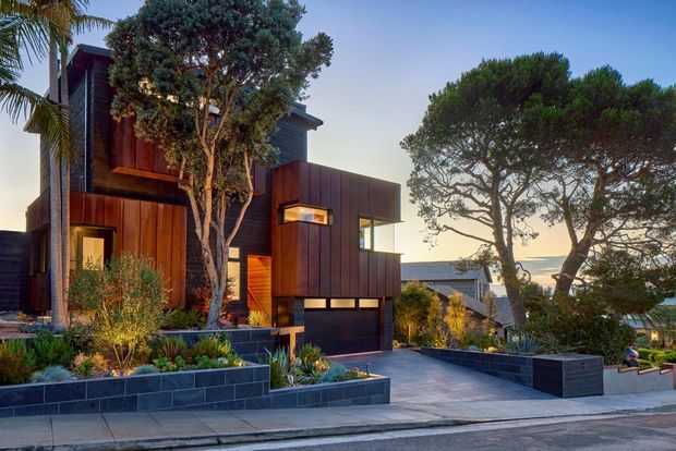 A contemporary multi-level residence viewed from the street at dusk, featuring dark timber cladding, projecting box-like volumes, large glazed openings, and an integrated garage. The home sits on a landscaped sloping site with layered dark masonry retaining walls, drought-tolerant planting, and a paved driveway. Mature sculptural trees partially screen the facade, with a distant coastal horizon visible beyond.