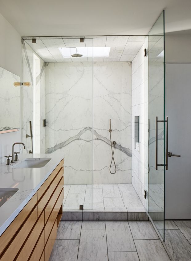 Bright contemporary bathroom with a large frameless glass walk-in shower, white marble-look wall and floor tiles with prominent grey veining, a floating timber vanity with white stone benchtop, and brushed metal tapware. A skylight and ceiling shower add natural light and function, while the palette is predominantly white, light grey, and warm timber.