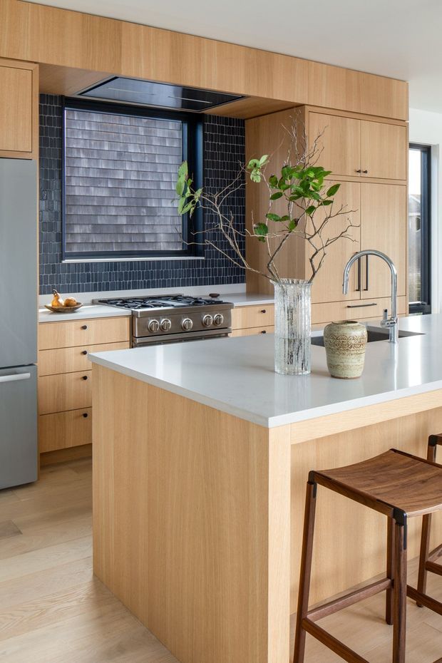 A contemporary kitchen featuring extensive light timber cabinetry, a large island with a pale stone benchtop and integrated seating, a stainless-steel range cooker, and a dark blue-black vertical tile splashback. A black-framed window, concealed rangehood, curved chrome tap, and decorative branches in a glass vase complete the bright, refined space.