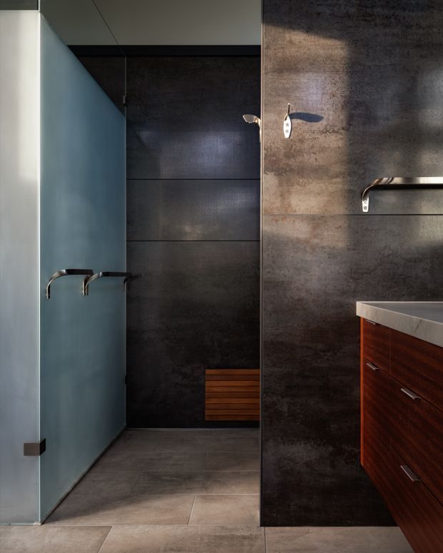 A compact, sophisticated bathroom with dark textured wall panels, large grey floor tiles, a frameless frosted-glass partition, minimalist chrome fixtures, and a rich reddish-brown timber vanity with a pale stone benchtop. The space has strong linear detailing and dramatic directional lighting.