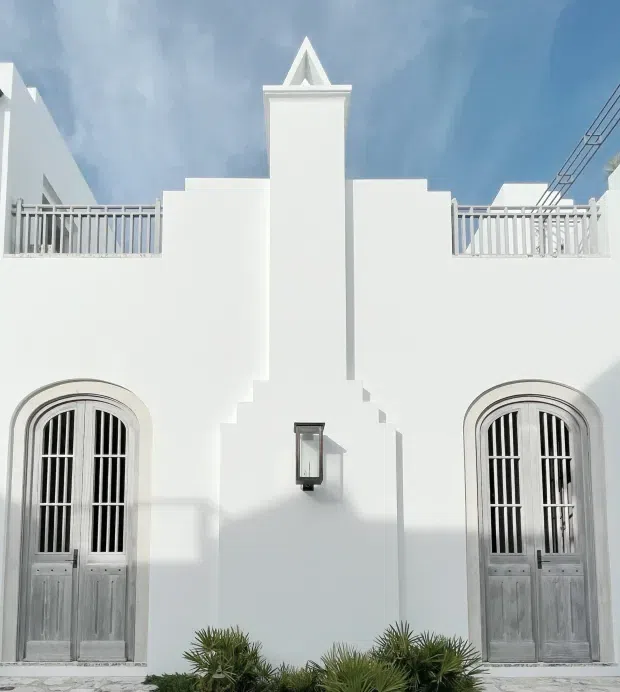 White symmetrical Mediterranean-style building facade with a central stepped tower and triangular decorative gable, rooftop terraces with white balustrades, and two tall arched grey timber doors with vertical metal bars. A wall-mounted lantern and low tropical landscaping frame the bright exterior.