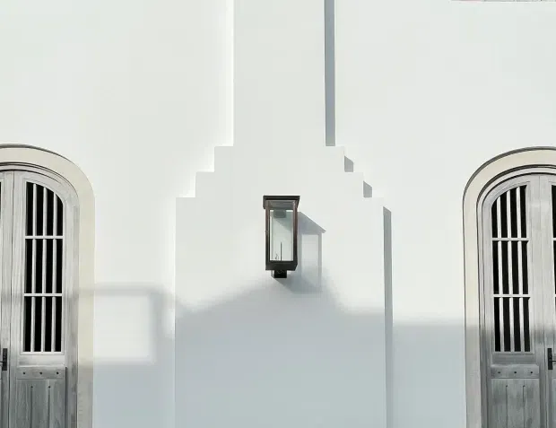 Symmetrical close-up of a white rendered facade featuring two tall arched timber doors with vertical black metal grilles and a small central wall-mounted lantern. Strong sunlight creates crisp architectural shadows, emphasizing the stepped parapet-like central form and decorative door surrounds.
