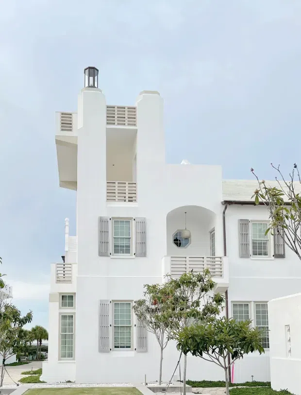 A bright white multi-storey residential facade with Mediterranean and coastal character. The building features a tall tower-like vertical element with a lantern-like rooftop window, open balconies with horizontal louvre balustrades, arched openings, white-framed multi-pane windows with grey shutters, and a pale pitched roof. Small landscaped trees and paved paths sit in front of the building under a pale blue sky.