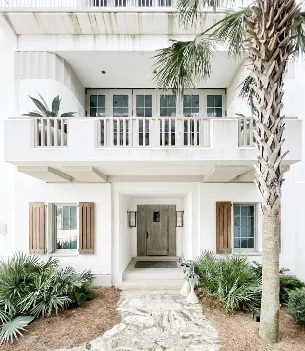 Front exterior view of a bright white two-storey coastal-style residence. The facade features a recessed entry with a weathered timber door, white vertical balustrades, an upper balcony, divided-light windows with timber shutters, exposed structural projections, and lush palm and drought-tolerant planting along a pale stone pathway.