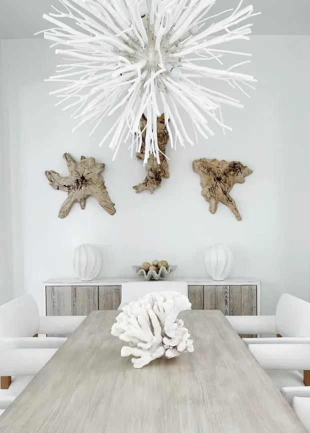 Bright, softly styled dining room with a long pale timber dining table surrounded by white upholstered seating. A large sculptural white pendant light hangs overhead, while the white walls feature natural driftwood-style wall sculptures. A weathered timber sideboard, white decorative vessels, and coastal organic accents complete the calm, light-toned interior.