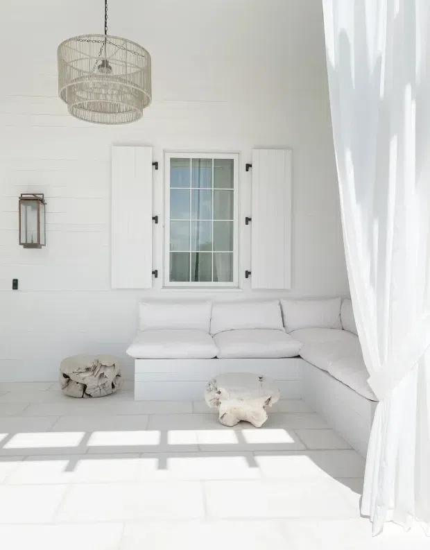 Bright white living room with horizontal white wall panelling, a large built-in L-shaped sectional beneath a shuttered multi-pane window, sheer floor-to-ceiling curtains, and sculptural pale wood or stone coffee tables. A woven pendant light and strong sunlight create an airy coastal atmosphere.