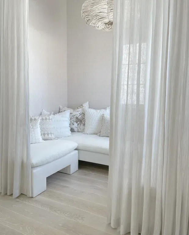 A softly lit, predominantly white living room viewed through sheer floor-to-ceiling curtains. The room contains a white L-shaped sectional sofa with textured and patterned cushions, pale timber flooring, light walls, and a sculptural woven pendant light. The curtains create a private, airy and serene atmosphere.