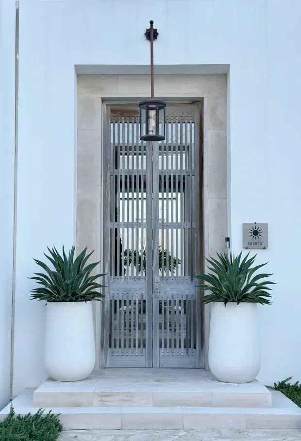 A symmetrical residential entrance with pale white walls, a large stone-framed doorway, and a grey timber slatted double door. A hanging lantern is centered above the entry, while two large white rounded planters with sculptural agave plants flank the steps. The entrance has a refined coastal appearance with light natural materials and strong vertical detailing.