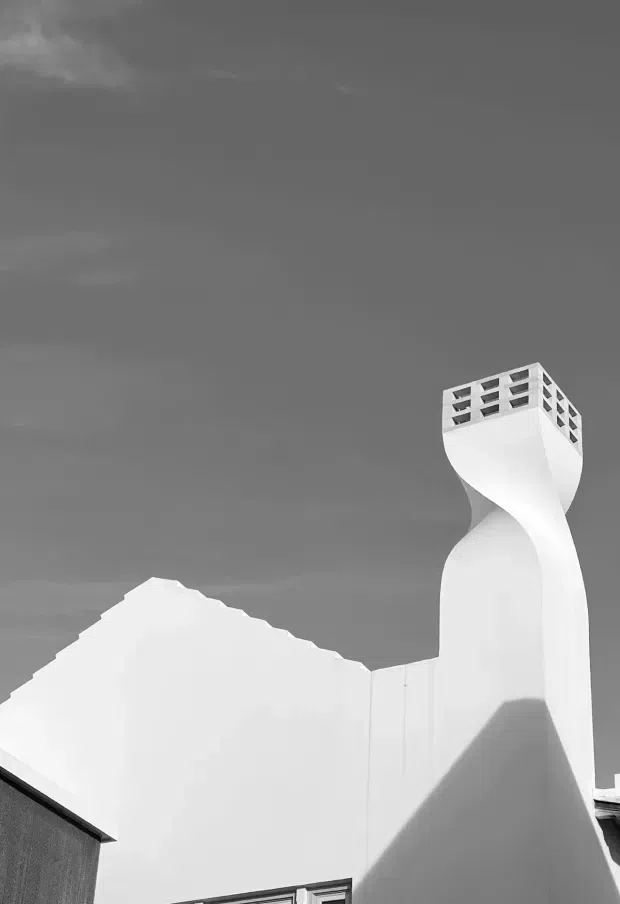 A monochrome exterior architectural photograph featuring a sculptural white rendered building facade and a tall, twisting chimney or ventilation tower capped with a perforated rectangular crown. A pitched roof and strong angular shadows are visible against an overcast sky. The image emphasises geometric form, smooth surfaces, and dramatic light and shadow.