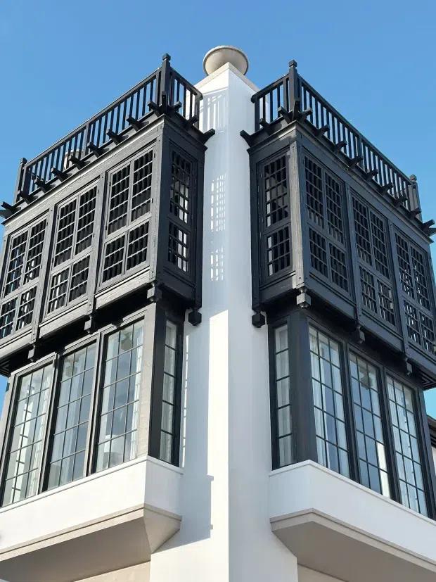 Upward-looking exterior view of a multi-storey building corner with a white rendered facade, large black-framed multi-pane windows, ornate dark projecting window screens or enclosed balconies, and black rooftop railings. A cylindrical rooftop feature is visible against a clear blue sky. The architecture has strong decorative detailing and a formal, historic-inspired character.