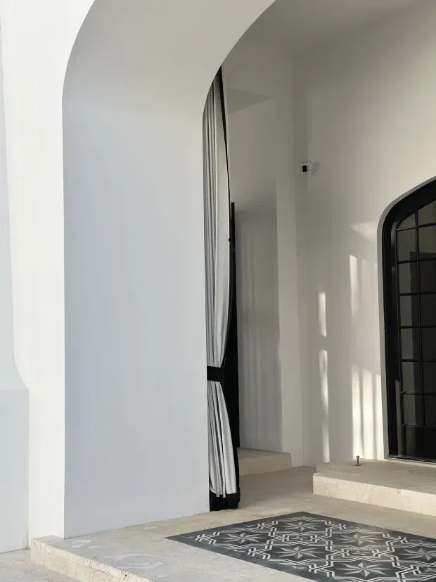 A bright, minimalist entryway with white plaster walls, a large curved archway, black-framed arched glazing or door, and full-height black-and-white curtains. Pale stone steps and flooring lead to a patterned monochrome rug, while sunlight creates strong vertical highlights on the walls.