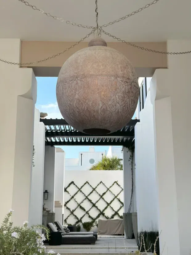 A bright Mediterranean-inspired outdoor courtyard or covered terrace framed by white stucco columns and walls. A large textured spherical pendant light hangs centrally from decorative chains, while a dark timber pergola shades a seating area. The courtyard includes white paving, trellis-patterned climbing greenery, planters, and low outdoor lounge furniture.