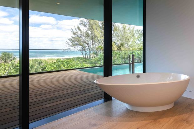 A contemporary bathroom featuring a freestanding oval bathtub on a warm timber floor. Full-height black-framed glazing opens to a timber deck and a turquoise pool, with a coastal ocean view beyond. The room has a clean, minimal composition and strong indoor–outdoor connection.