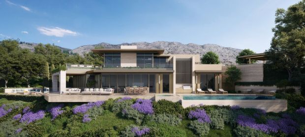 A wide exterior view of a contemporary multi-level residence set into a landscaped hillside, with broad glazing, horizontal roof planes, deep outdoor terraces, a long swimming pool, and a mountain backdrop. The garden is planted with dense low greenery and vivid purple flowering shrubs.