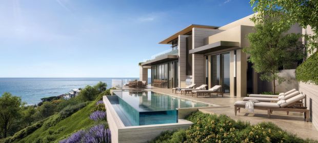 A contemporary multi-level coastal residence perched above the ocean, featuring expansive glazing, projecting flat roof planes, a large infinity-edge swimming pool, landscaped terraces, and outdoor lounge areas. The architecture uses clean geometric forms and neutral-toned finishes, with ocean views and landscaped grounds dominating the setting.