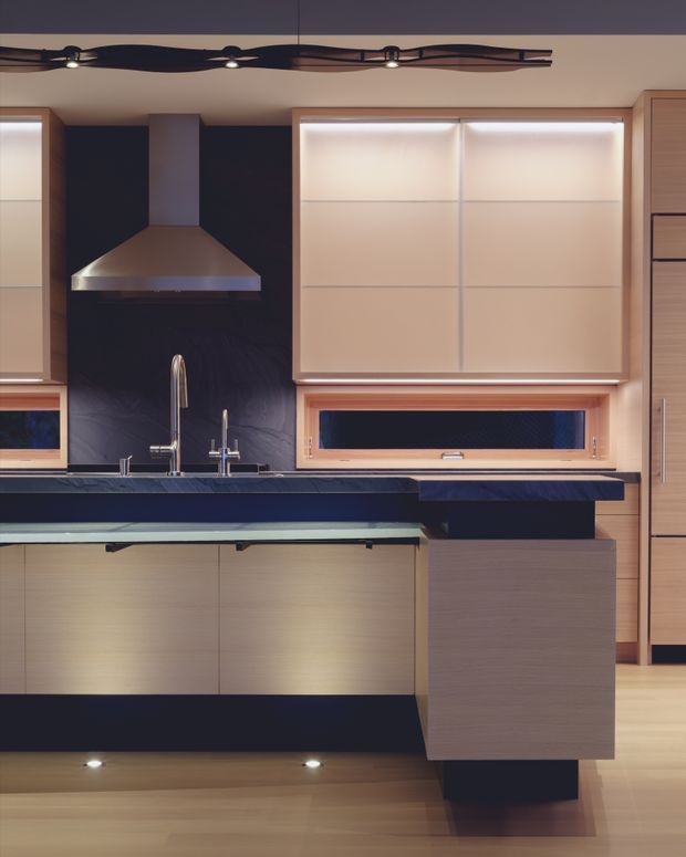 A contemporary kitchen interior with a central island, dark stone benchtops, pale timber cabinetry, a stainless-steel rangehood, integrated sink and tapware, and large windows fitted with translucent blinds. Linear concealed lighting and dark feature surfaces create a refined, high-contrast composition.
