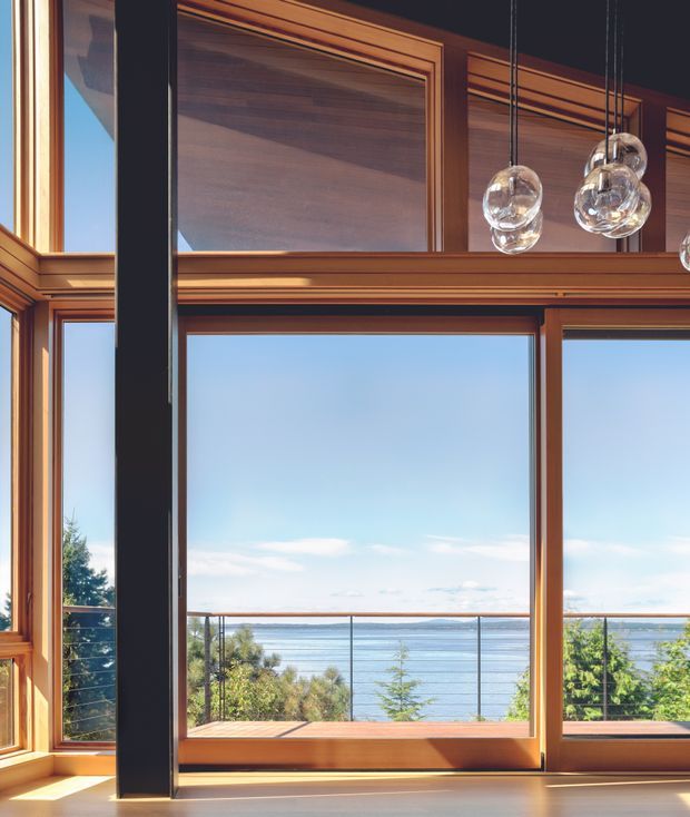 Interior architectural view dominated by extensive warm timber-framed glazing overlooking a body of water and landscaped vegetation. A dark structural post divides the composition, while a high-level timber-lined section and several clear glass globe pendant lights add detail. The space appears bright and airy, with a strong indoor-outdoor connection and a coastal or waterfront outlook.