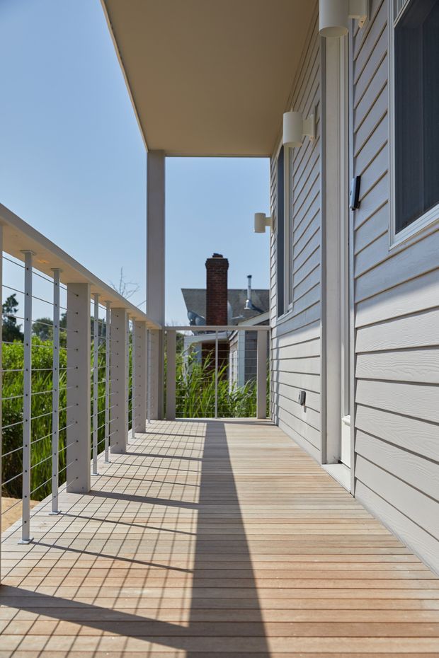 A bright outdoor elevated walkway or balcony running alongside a contemporary light-clad house. The deck has narrow timber boards, white horizontal weatherboard cladding, slim white posts, and cable balustrading. Strong sunlight casts geometric shadows across the deck, with neighbouring residential buildings and vegetation visible beyond.
