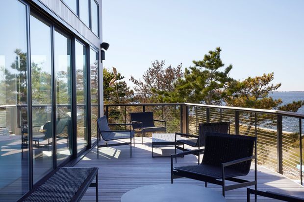 A contemporary waterfront-facing outdoor terrace or balcony with dark grey decking, black metal-framed full-height glazing, and a horizontal metal balustrade. Black outdoor lounge chairs and a low coffee table are arranged across the spacious deck, with trees and water visible beyond. The scene is bright, airy, and minimalist with strong indoor-outdoor connection.