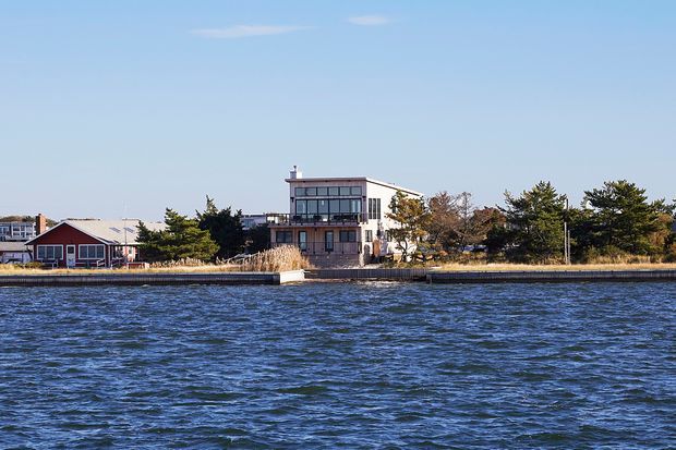 Exterior view across a body of water toward a waterfront property. The main subject is a contemporary multi-level house with extensive glazing, a flat roof, balconies or terraces, and light-coloured walls, accompanied by a smaller red gabled building and mature trees along the shoreline.