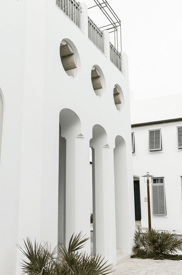 A bright white architectural courtyard or exterior walkway framed by a tall facade with repeated pointed arches and circular recessed openings. The upper level has grey metal balustrades and a lightweight pergola-like frame, while a neighbouring white building with louvred windows is visible beyond. Pale stone paving and sculptural drought-tolerant planting complete the scene.
