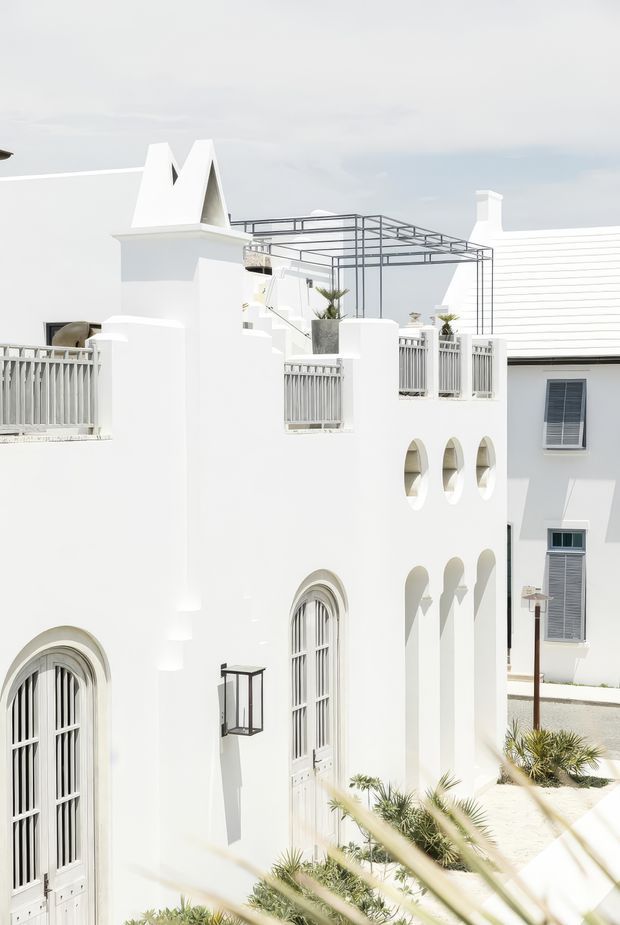 Bright exterior view of a white, multi-level Mediterranean-influenced building with arched windows and openings, decorative parapets, rooftop terraces, pale grey railings, and a lightweight metal pergola structure. Palm-like landscaping and adjacent white buildings reinforce a sunlit coastal resort or residential setting.