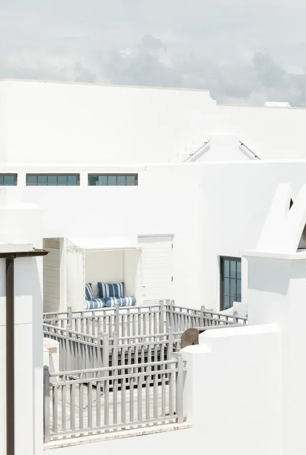 Bright exterior view of a white multi-level building with clean geometric forms, narrow dark-framed windows, rooftop structures, and a small elevated terrace or balcony enclosed by pale grey railings. The terrace includes a built-in or freestanding daybed with blue-and-white striped cushions. The scene has a strong white coastal aesthetic and abundant daylight.