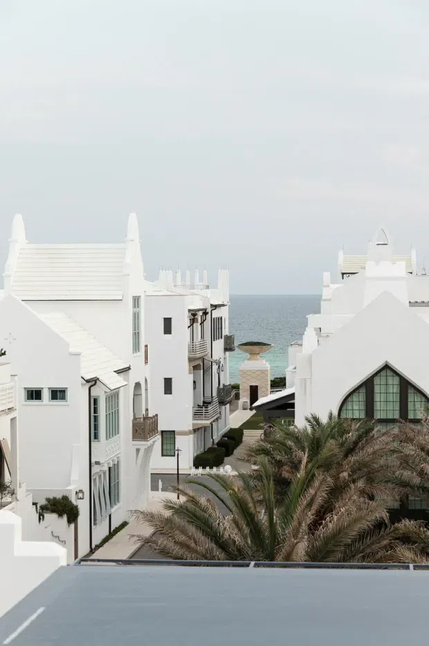 Elevated exterior view down a whitewashed coastal residential development toward the ocean. The buildings have crisp stucco facades, steep and varied roof forms, balconies, arched and gridded windows, and a Mediterranean or resort-like character. Palm trees occupy the foreground, with a paved pedestrian lane and landscaped hedges between the buildings.