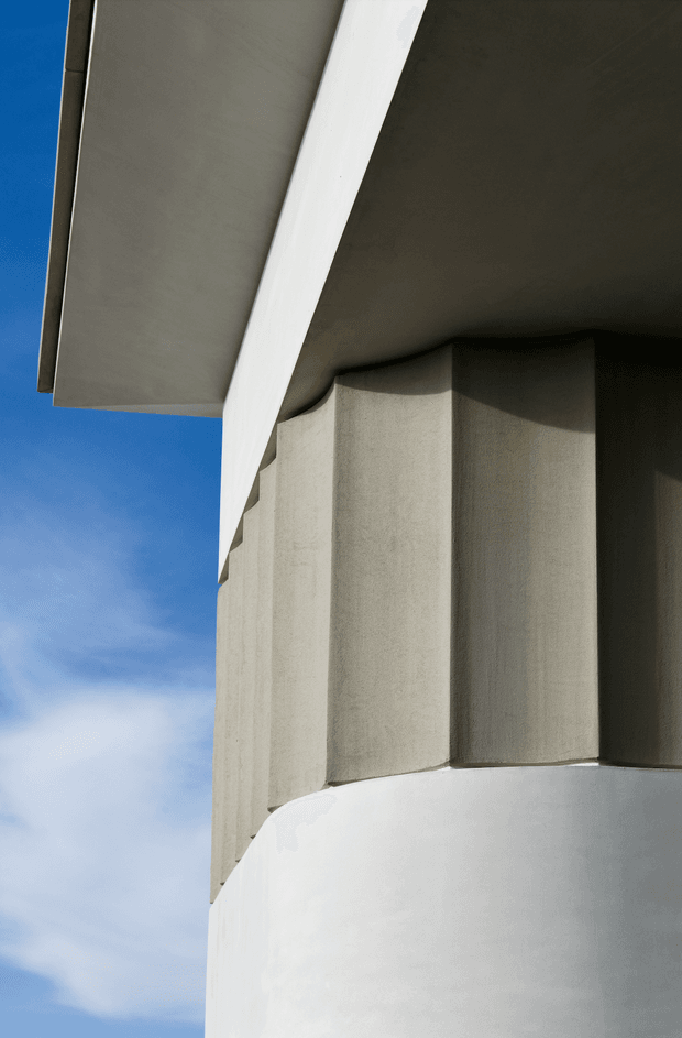 Close-up architectural exterior detail showing a sculptural curved white rendered wall or parapet beneath a projecting roof or canopy, with tall faceted concrete or plaster wall elements and deep shadow lines. The composition emphasizes geometric massing, smooth and textured surfaces, and a bright blue sky.