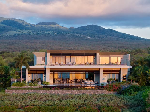 A large modern two-storey residence set within landscaped grounds, with extensive floor-to-ceiling glazing, broad horizontal overhangs, open terraces, and warm interior lighting. The house sits against a wooded mountainous backdrop and is surrounded by layered garden planting.