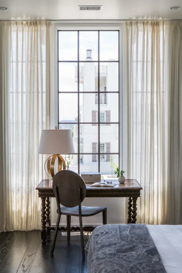 A bright, refined bedroom corner featuring a small writing desk positioned in front of a tall, divided-pane window. Sheer floor-to-ceiling curtains frame the window, while an ornate dark wood desk with turned legs, a sculptural table lamp, a simple chair, books, glasses, and a small vase create a compact workspace. Part of the bed with a grey patterned cover is visible in the foreground. The room has dark timber flooring, pale walls and ceiling, and a light-filled traditional interior character.
