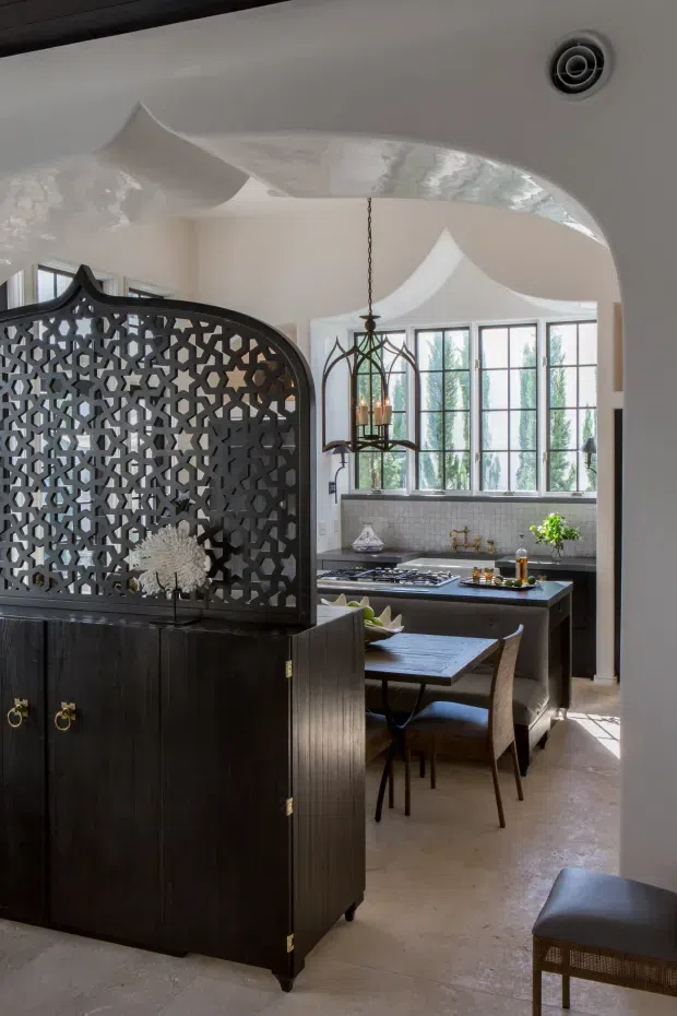 A refined kitchen and dining area viewed through an arched opening. The room features a dark timber cabinet with an ornate geometric screen, dark kitchen island and cabinetry, a pale tiled backsplash, expansive black-framed windows, and a sculptural plaster ceiling with curved forms. A decorative hanging lantern and compact dining table complete the space.