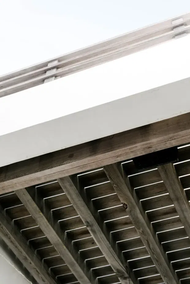 Upward-looking architectural detail showing exposed weathered timber rafters and decking beneath a projecting roof or pergola, with a clean white structural fascia and pale linear elements above. The image is an abstract close-up of building structure rather than a complete room or facade.
