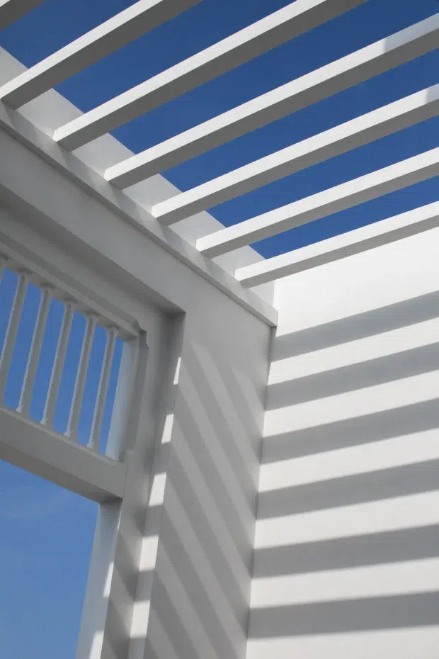 Upward architectural detail of a bright white exterior or semi-outdoor structure, featuring evenly spaced roof beams forming a pergola-like canopy, a white balustrade, and strong linear shadows cast across the walls. Clear blue sky is visible through the open roof and railing, creating a crisp, geometric composition.