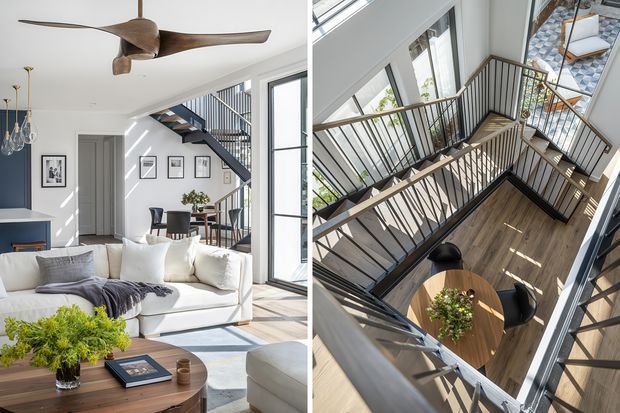 Split-view interior photograph showing a bright contemporary living room and a dramatic multi-level stair and landing void. The living area has a large white sectional sofa, timber coffee table, framed wall art, pendant lights, full-height black-framed glazing, and a sculptural timber ceiling fan. The adjacent view looks down over dark metal balustrades, timber flooring, stairs, and a round dining table in the lower-level space, with strong natural light entering from roof glazing.