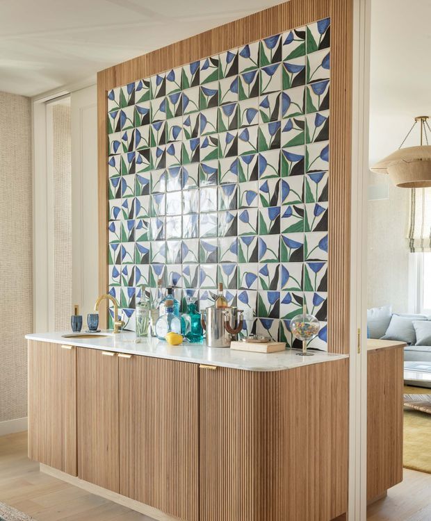 A bright, carefully detailed open-plan interior featuring a built-in fluted timber bar or kitchen cabinet with a pale stone benchtop and an expansive geometric blue, green and white tiled feature wall. A brass-toned tap, glassware and bottles sit on the counter, while a softly furnished living area is visible beyond the opening. Vertical timber battens, patterned wall tiles, light walls and natural daylight create a refined yet playful atmosphere.