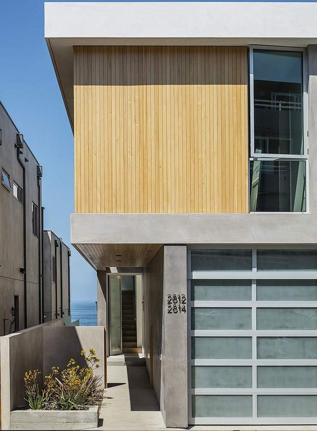 Front exterior view of a contemporary multi-storey residence on a narrow urban lot. The facade combines pale concrete, a large vertical timber-clad upper wall, dark-framed glazing, and a frosted-glass sectional garage door. A recessed entry walkway runs beside the garage, with a glimpse of blue water visible between neighbouring buildings.