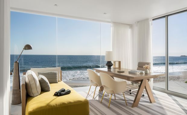 Bright contemporary open-plan living and dining area with floor-to-ceiling glazing overlooking a sandy beach and ocean. The room features a mustard-yellow sofa, neutral dining table and sculptural chairs, sheer white curtains, pale flooring, and restrained modern furnishings.