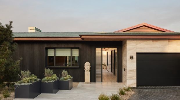 Single-storey contemporary residence viewed from the front, with dark vertical cladding, pale stone garage surround, low-pitched roof forms, black garage door, and a sheltered entry opening onto a light-toned interior corridor. Minimal landscaped planters and paving frame the facade.